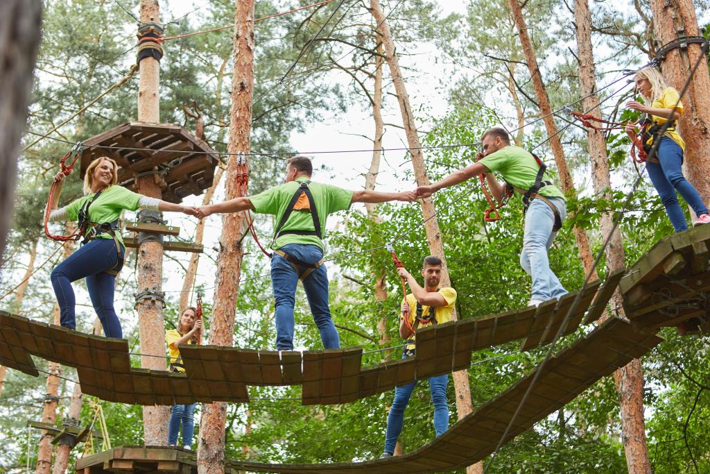 Teammitglieder halten sich gemeinsam auf einem Klettergarten an den Händen und überqueren eine schwingende Holzbrücke Teammitglieder halten sich gemeinsam auf einem Klettergarten an den Händen und überqueren eine schwingende Holzbrücke
